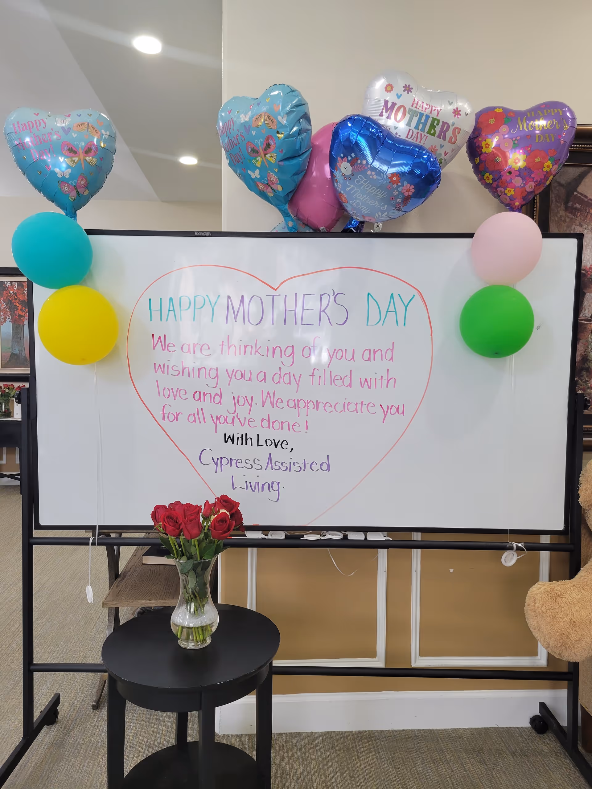 A whiteboard decorated with colorful balloons and a handwritten message inside a large heart shape that reads: 'HAPPY MOTHER'S DAY We are thinking of you and wishing you a day filled with love and joy. We appreciate you for all you've done! With Love, Cypress Assisted Living.' Below the whiteboard is a small black table with a vase of red roses. The setting appears to be an indoor common area.