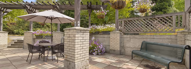 Shaded outdoor patio with a table and umbrella, pergola, hanging flower baskets, brick planters, and a bench.