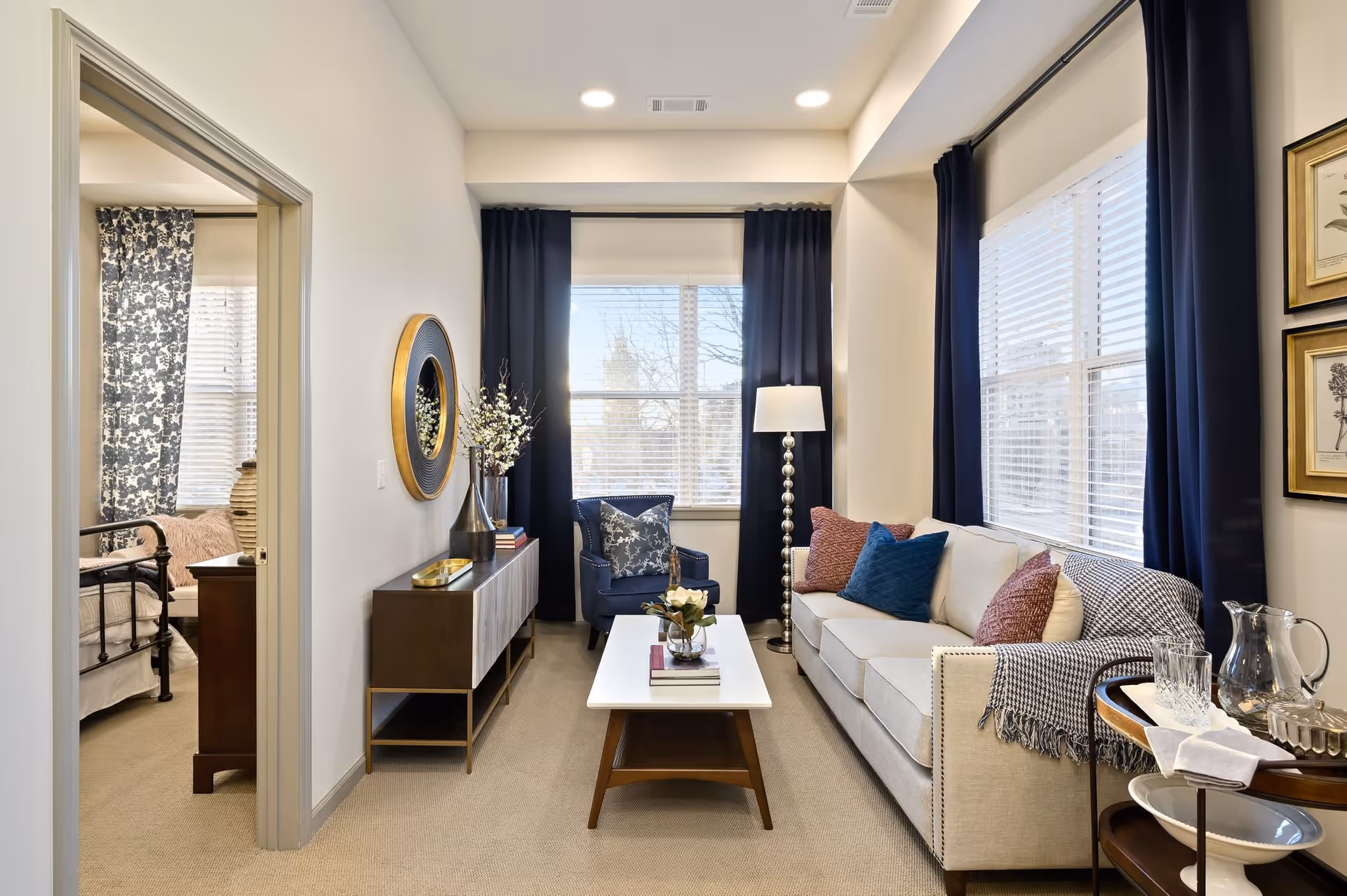 A bright and cozy living room with a beige sofa adorned with colorful pillows and a throw blanket, a blue armchair, a white coffee table with books and a flower vase, a floor lamp, and dark blue curtains framing two large windows. To the left, an open doorway reveals a bedroom with a bed, nightstand, and patterned curtains.