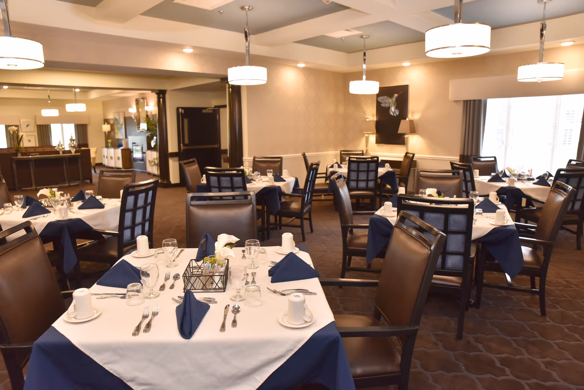 Dining room with multiple tables set with white tablecloths, navy napkins, place settings and chairs under pendant lights.