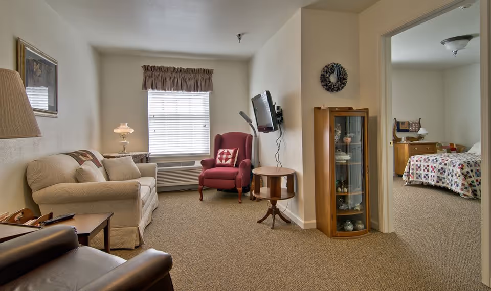 Cozy living room with a sofa, armchair, wall-mounted TV, small tables and a display cabinet, with a bedroom visible through a doorway.