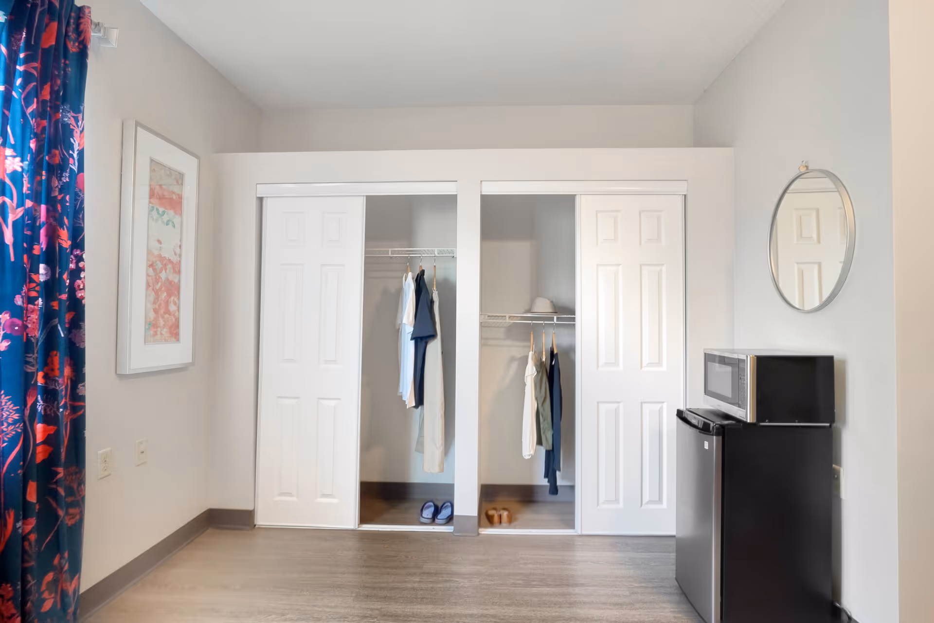 Interior view of a room with two open closets containing hanging clothes and shoes. To the right, there is a small black refrigerator with a microwave on top and a round mirror hanging on the wall above it. On the left side, there is a window with blue and red floral curtains and a framed artwork on the wall.