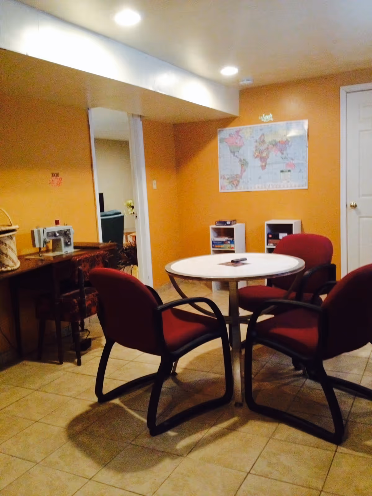 A small room with orange walls and tiled floor featuring a round white table surrounded by four red cushioned chairs with black frames. On the left side, there is a wooden desk with a sewing machine and a basket on top, and a chair with a patterned cushion. A world map is mounted on the wall above two small white shelves holding board games. A white door is visible on the right side of the room.