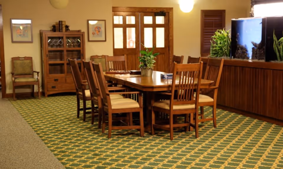 A dining room with a large wooden table surrounded by eight wooden chairs with cushioned seats. The room has a green patterned carpet and a wooden cabinet with glass doors against the wall. There are framed pictures on the wall and a large fish tank on the right side. A potted plant is placed in the center of the table.