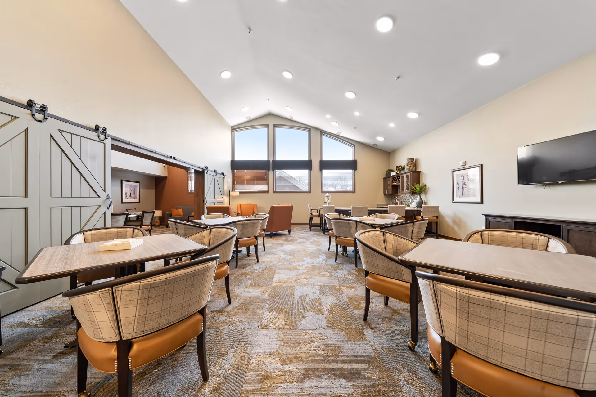 A bright and spacious dining room with multiple tables and cushioned chairs arranged neatly. The room features high vaulted ceilings with recessed lighting, large windows letting in natural light, and a neutral color palette. There is a TV mounted on the wall and a cabinet with decorative items on the right side.