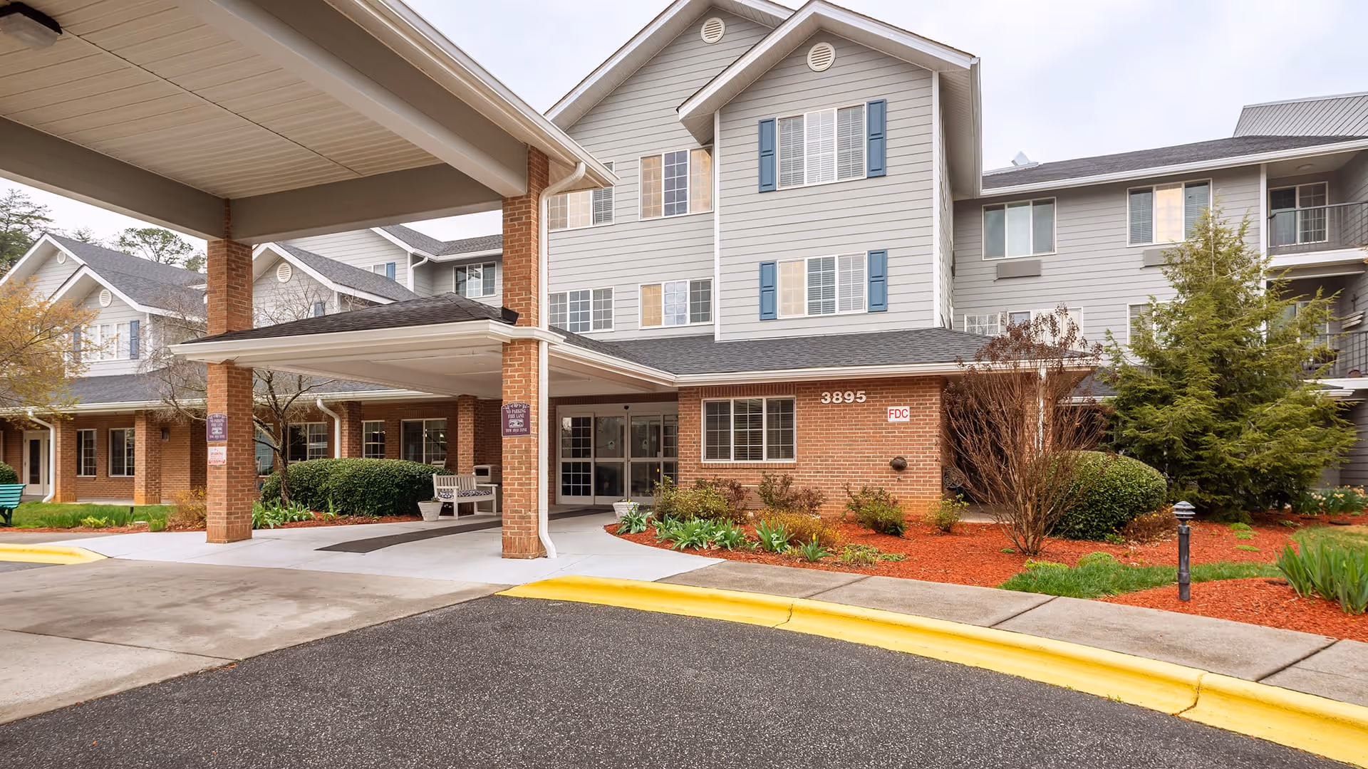 Exterior view of Creekside Terrace senior living facility showing a covered entrance with brick pillars, a driveway with yellow curb, landscaped bushes and trees, and a multi-story building with gray siding and blue shutters.