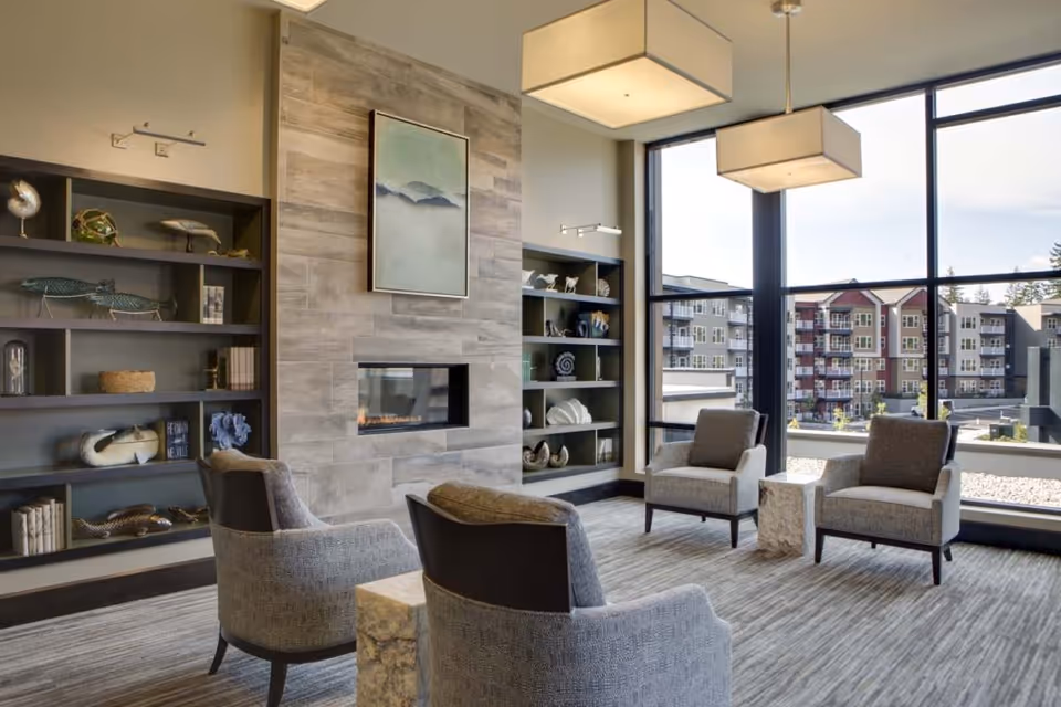 A modern living room with large floor-to-ceiling windows showing an exterior view of apartment buildings. The room features a tiled fireplace with a painting above it, built-in shelves with decorative items, four upholstered armchairs arranged around two small stone side tables, and two rectangular pendant lights hanging from the ceiling.