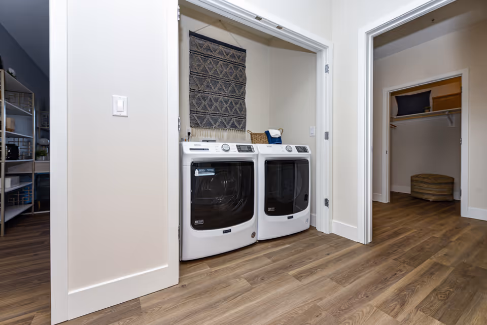 A laundry area with a white front-loading washing machine and dryer side by side in a recessed nook. Above the machines is a decorative woven wall hanging. The floor is wood with a natural finish, and adjacent rooms with open doors are visible, including a closet with shelves and storage items.