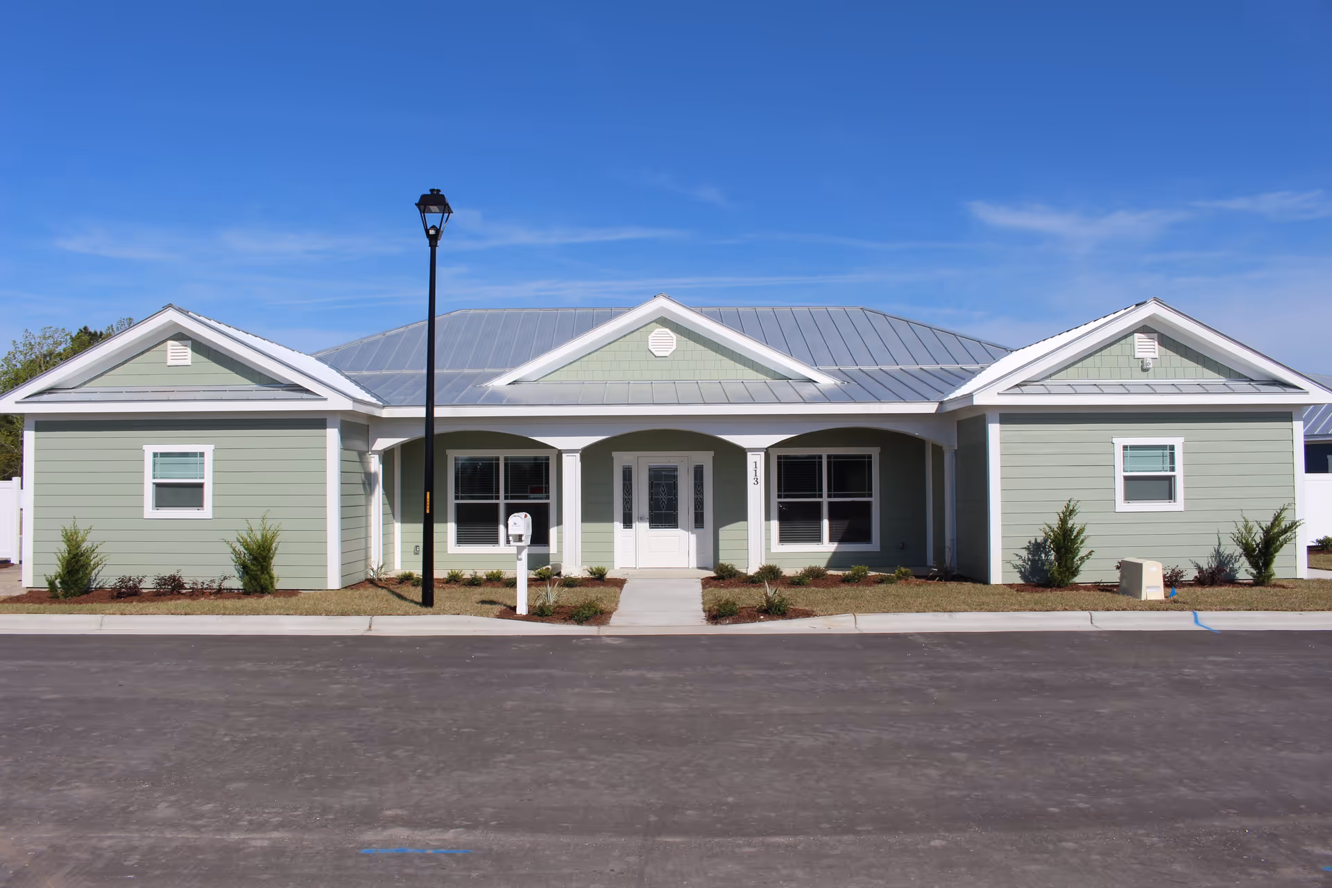 Front exterior view of a single-story residential building with light green siding, white trim, and a metal roof under a clear blue sky. The building has a central entrance with a white door and two large windows on either side. There is a black street lamp and a white mailbox in front of the building, along with small shrubs and landscaping.