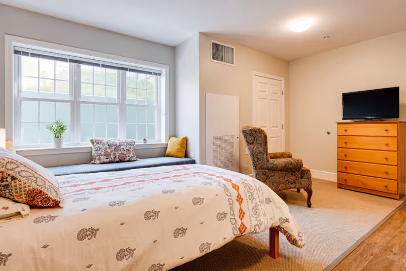 A bright bedroom with a large window featuring a cushioned window seat with pillows. The bed has a patterned white and orange bedspread with matching pillows. There is a floral upholstered armchair near a wooden dresser with a flat-screen TV on top. The room has beige walls and carpeted flooring with a wooden floor border.