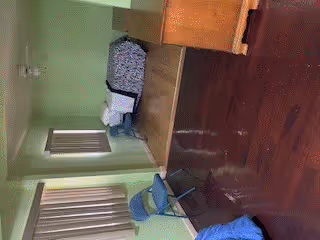 A simple bedroom with light green walls and wooden flooring. The room contains a single bed with a patterned blanket, a small table with a blue chair, and a window with vertical blinds letting in natural light.