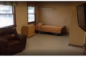 A simple room with a single bed covered with a brown blanket, a wooden dresser with two windows above it, a brown leather armchair, and a wall-mounted television on the right side.