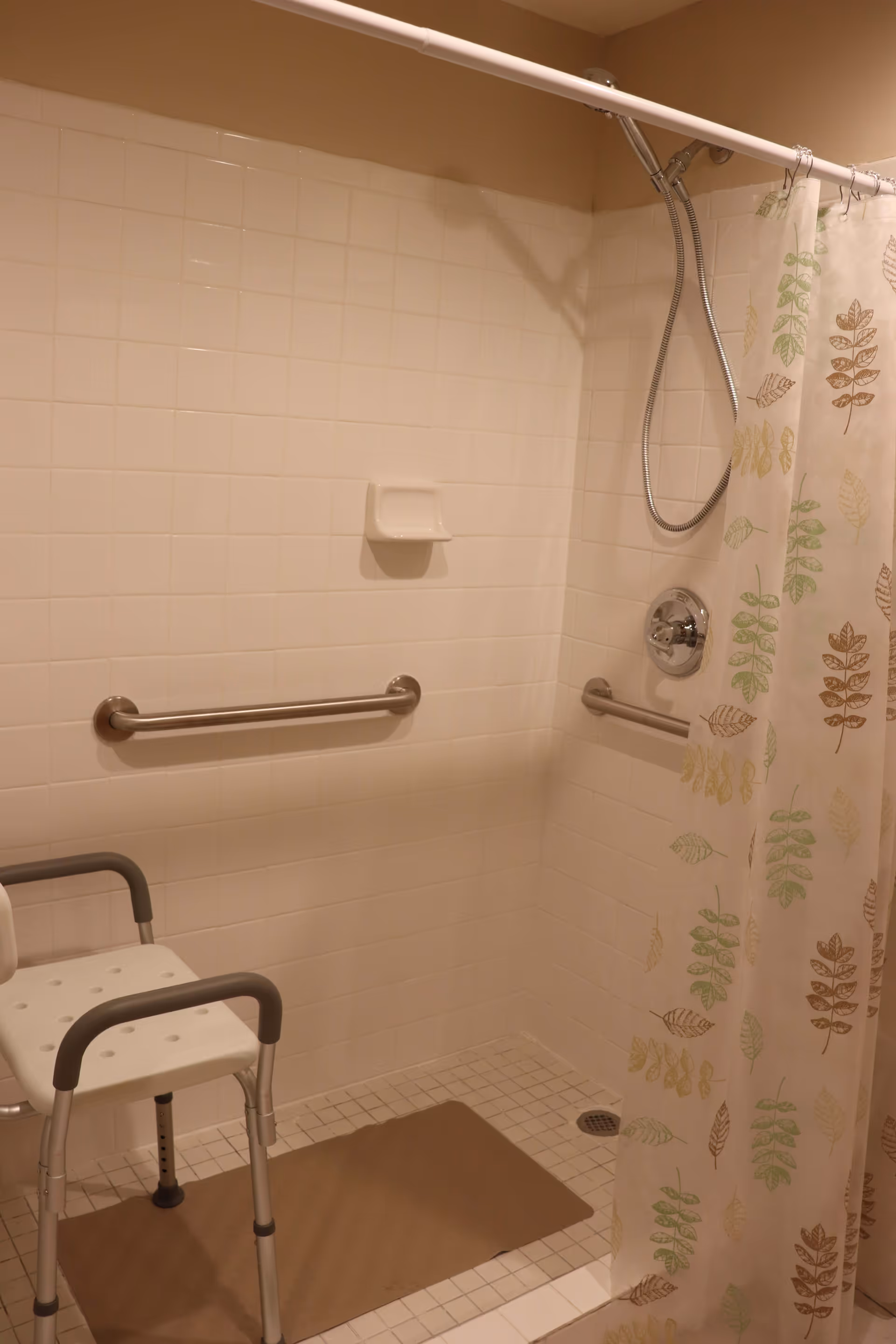 Accessible tiled shower with grab bars, a shower chair, handheld showerhead, and a patterned shower curtain.