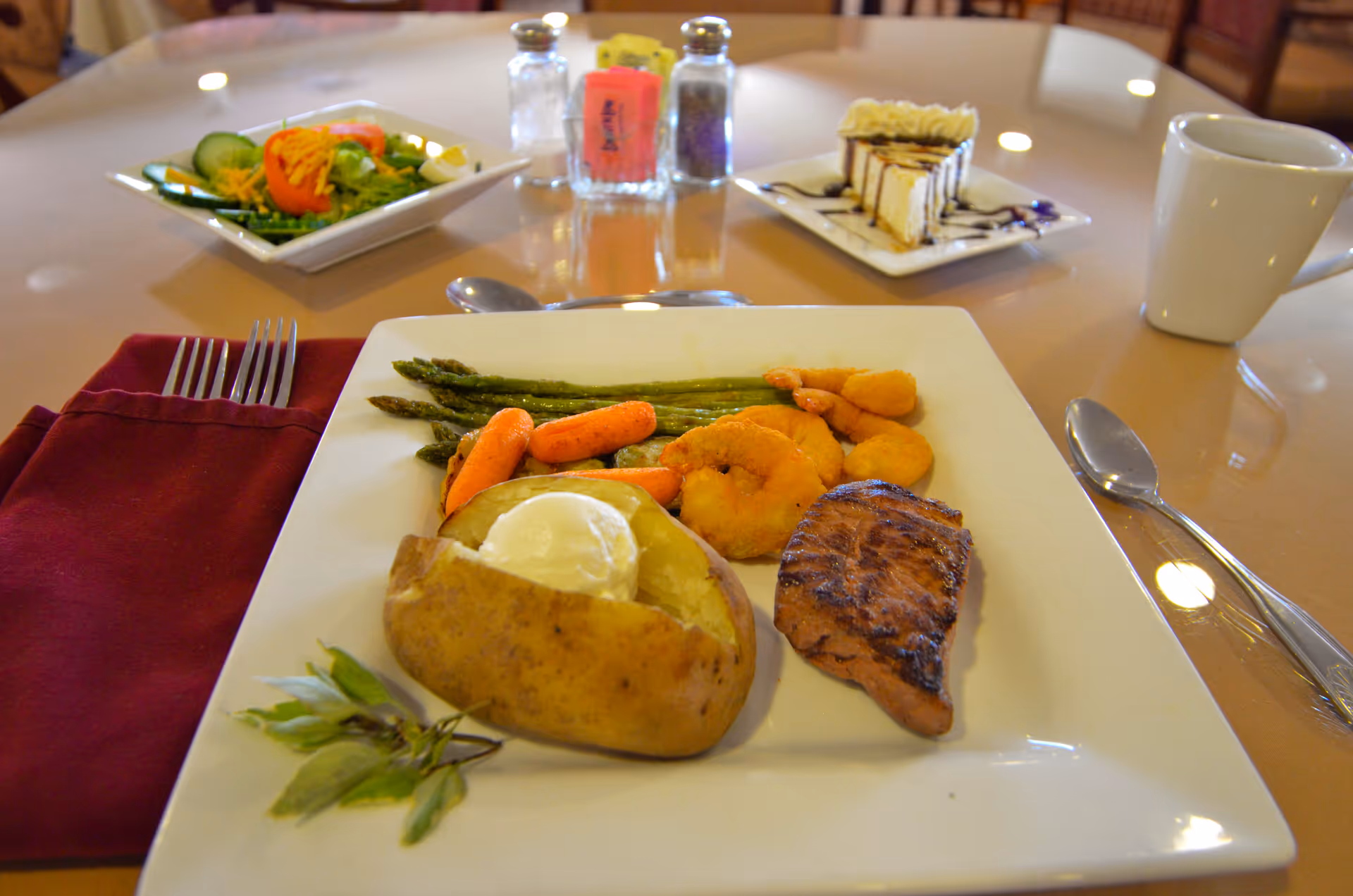 A plated meal on a square white plate featuring a baked potato with sour cream, grilled steak, fried shrimp, steamed asparagus, and baby carrots. In the background, there is a side salad with cucumbers, tomatoes, and shredded cheese, a slice of layered dessert with chocolate drizzle, salt and pepper shakers, and a white coffee mug on a round table with a maroon napkin and silverware.