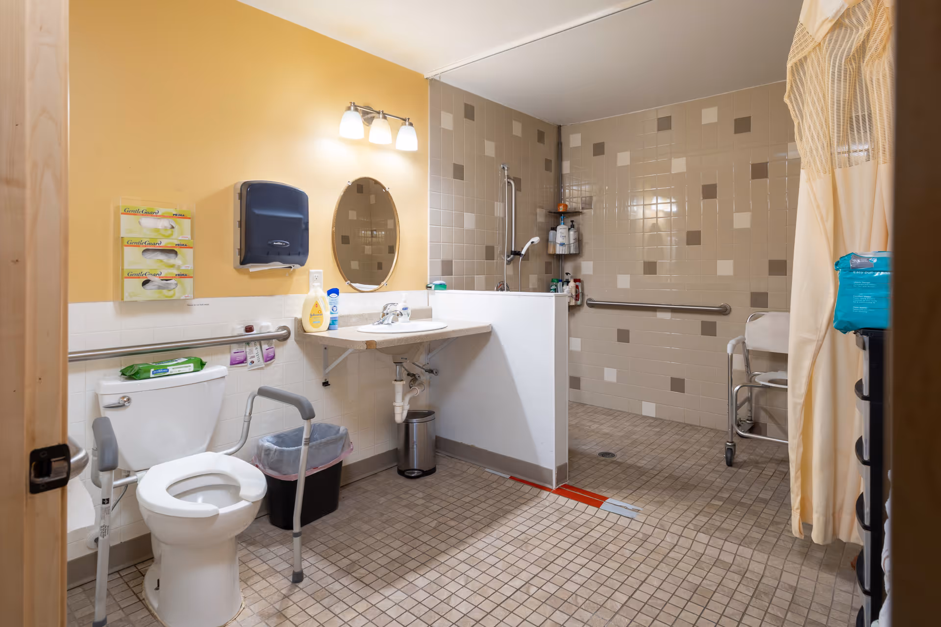 A spacious, accessible bathroom with beige and white tiled walls and floor. The room features a toilet with support rails, a sink with a mirror and lighting above, a trash bin, and a walk-in shower area with grab bars and a shower chair. Various hygiene products and paper towel dispensers are mounted on the walls.