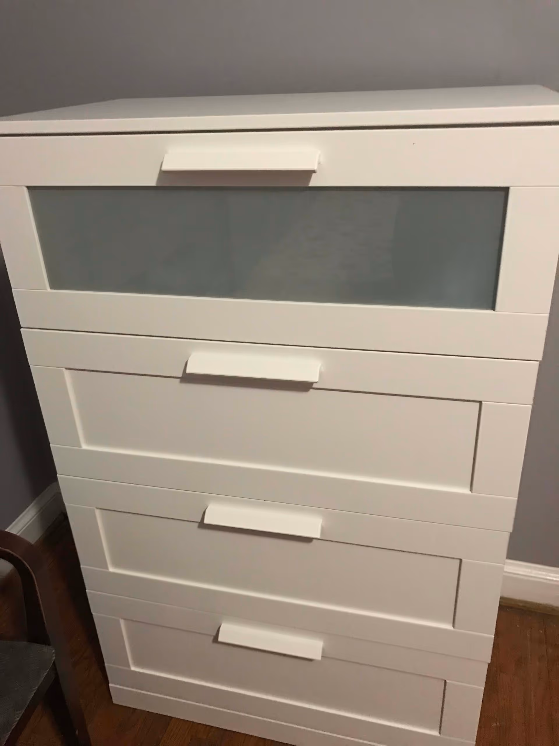 A white chest of drawers with four drawers, the top drawer has a frosted glass panel, placed against a gray wall on a wooden floor.
