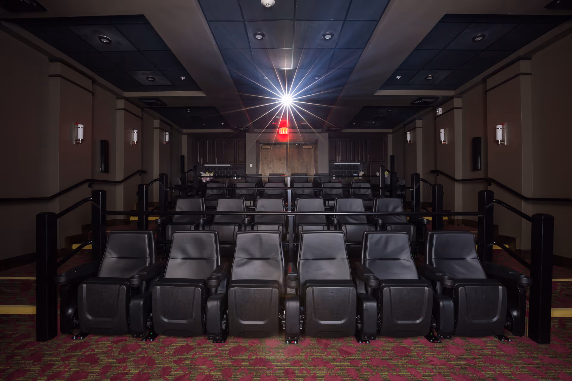 Small private movie theater with rows of black reclining seats, a projector beam and an illuminated exit sign.