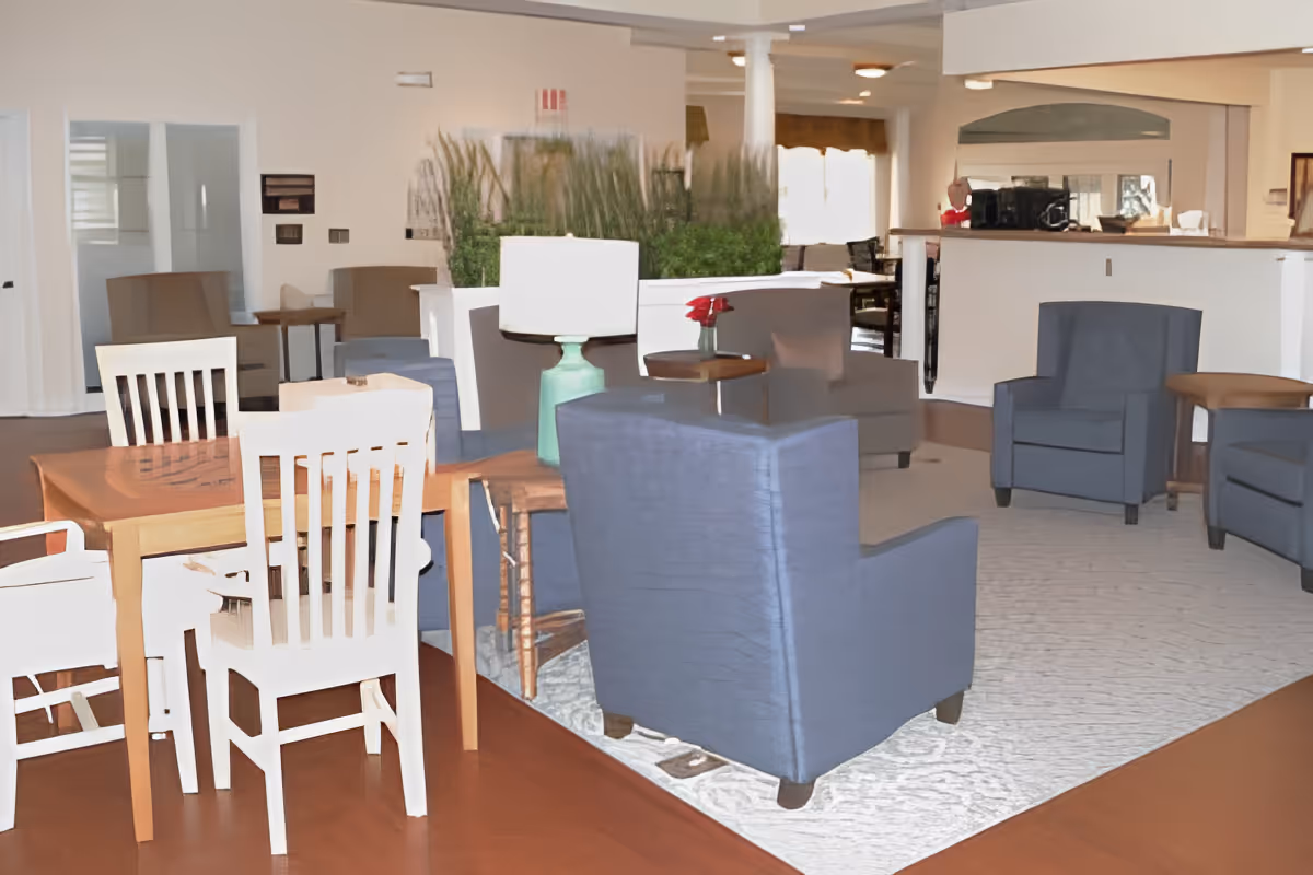 Open senior living common area with a wooden dining table and chairs, blue upholstered armchairs on a rug, and a reception counter in the background.