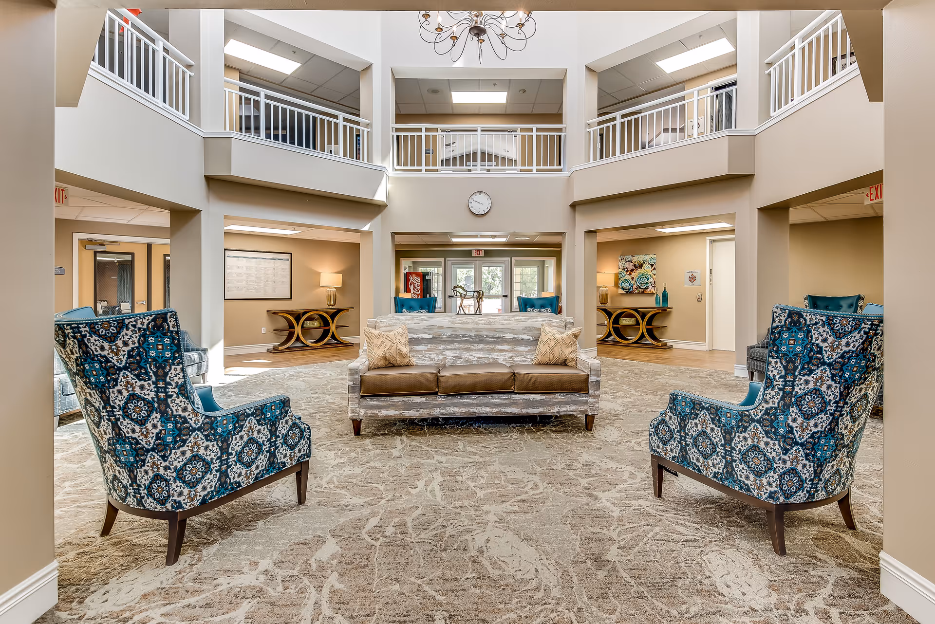 Bright two-story senior living lobby with patterned armchairs, a central sofa, and balcony railings overlooking the seating area.