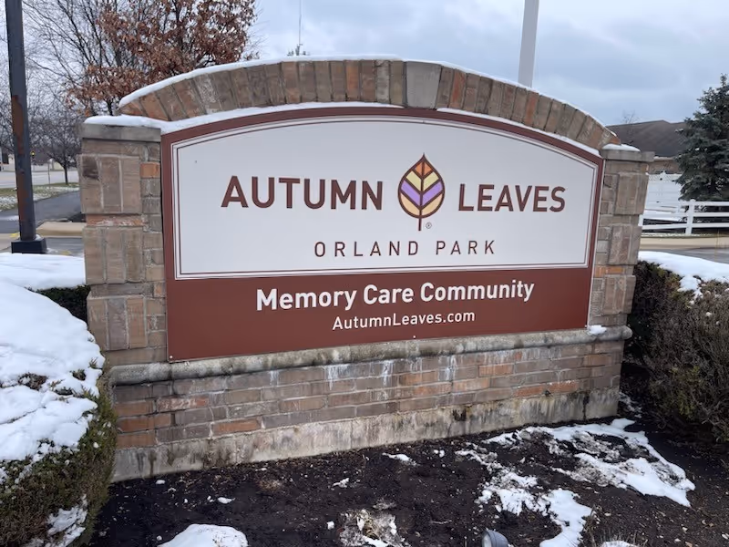 Outdoor sign for Autumn Leaves Orland Park Memory Care Community with a logo of a stylized leaf, surrounded by snow-covered ground and bushes.