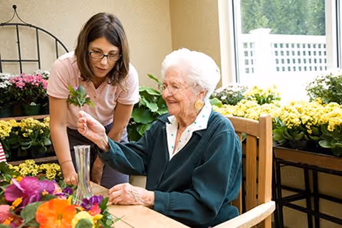 An elderly woman arranging flowers at a table while a caregiver assists her in a bright room filled with potted plants.