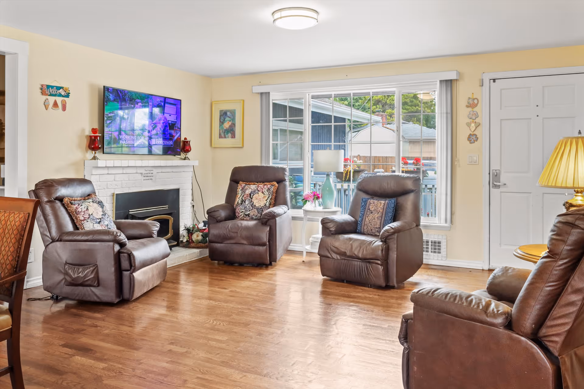 A cozy living room with four brown leather recliners arranged around a white brick fireplace with a mounted flat-screen TV above it. The room has hardwood floors, a large window with a view of the outside, a small white side table with a lamp and flowers, and a white front door. The walls are painted light yellow and decorated with framed artwork and small hanging decorations.