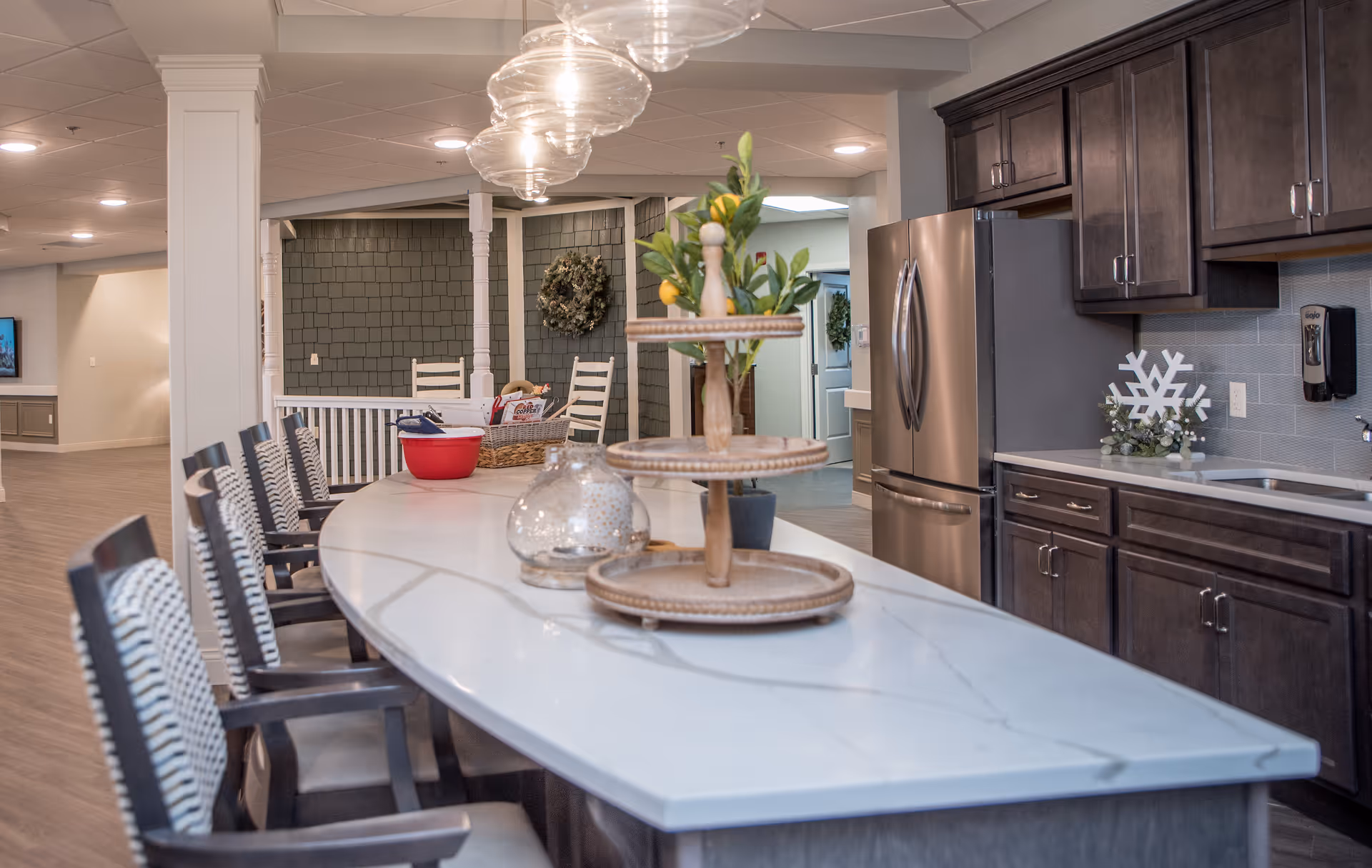 Open communal kitchen and dining area with a long marble-topped island, seating, pendant lights, and dark wood cabinetry.