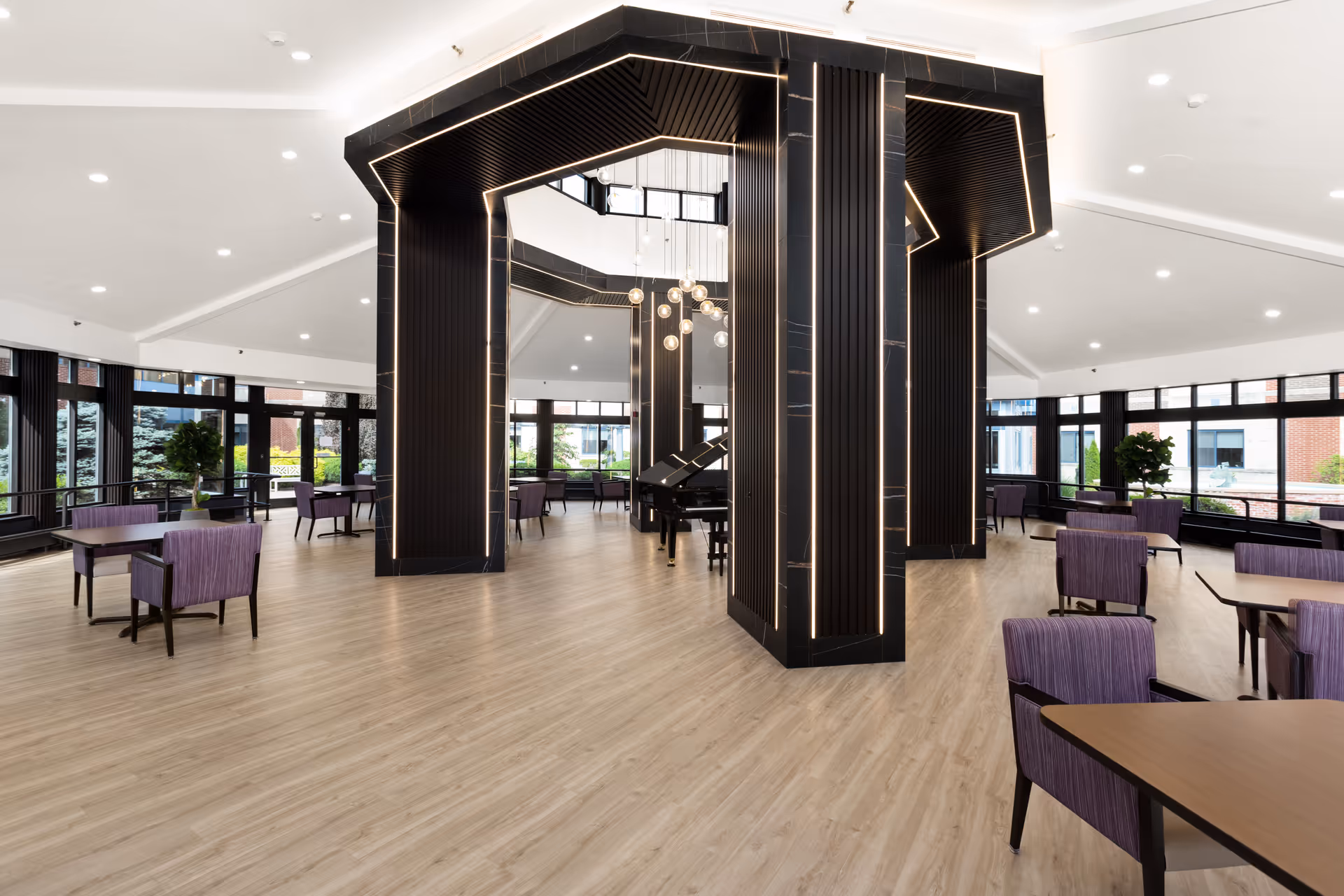 Spacious, modern common area with light wood flooring, large black pillars with integrated lighting, multiple tables with purple upholstered chairs, large windows letting in natural light, and a grand piano in the center under a contemporary chandelier.