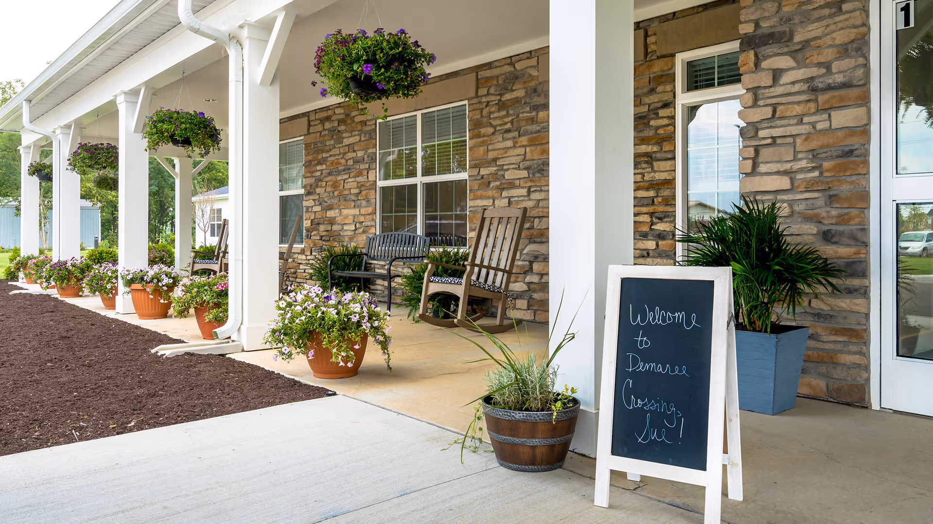 Outdoor covered patio area at Demaree Crossing with hanging flower baskets, potted plants, a bench, rocking chairs, and a chalkboard sign that reads 'Welcome to Demaree Crossing, Inc!'