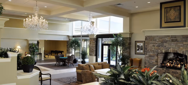 Bright communal lounge with chandeliers, comfortable seating, a stone fireplace, plants, and a piano near the entrance.