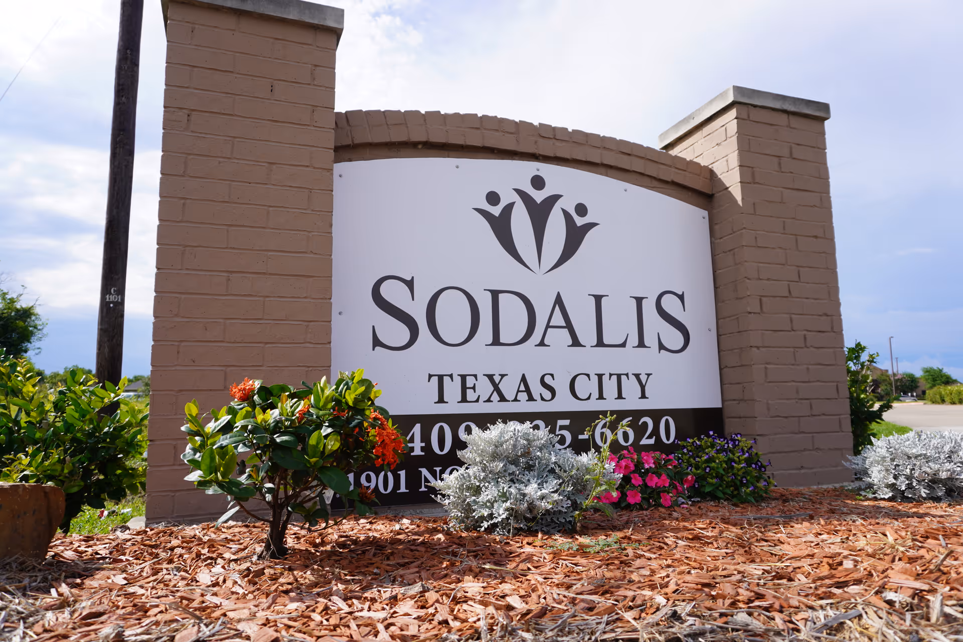 A large brick sign for Sodalis Texas City surrounded by small flowering plants and mulch, with a partly cloudy sky in the background.