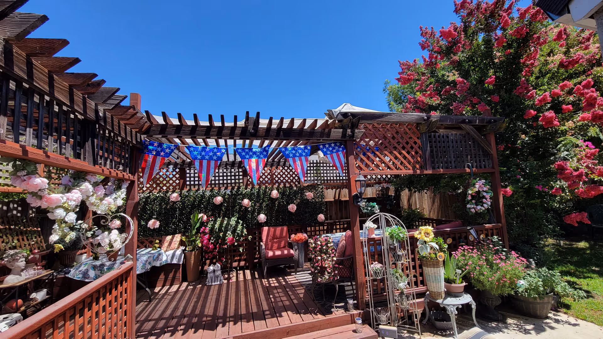 Sunlit wooden pergola-covered deck with seating, potted flowers, and American flag bunting in a garden.