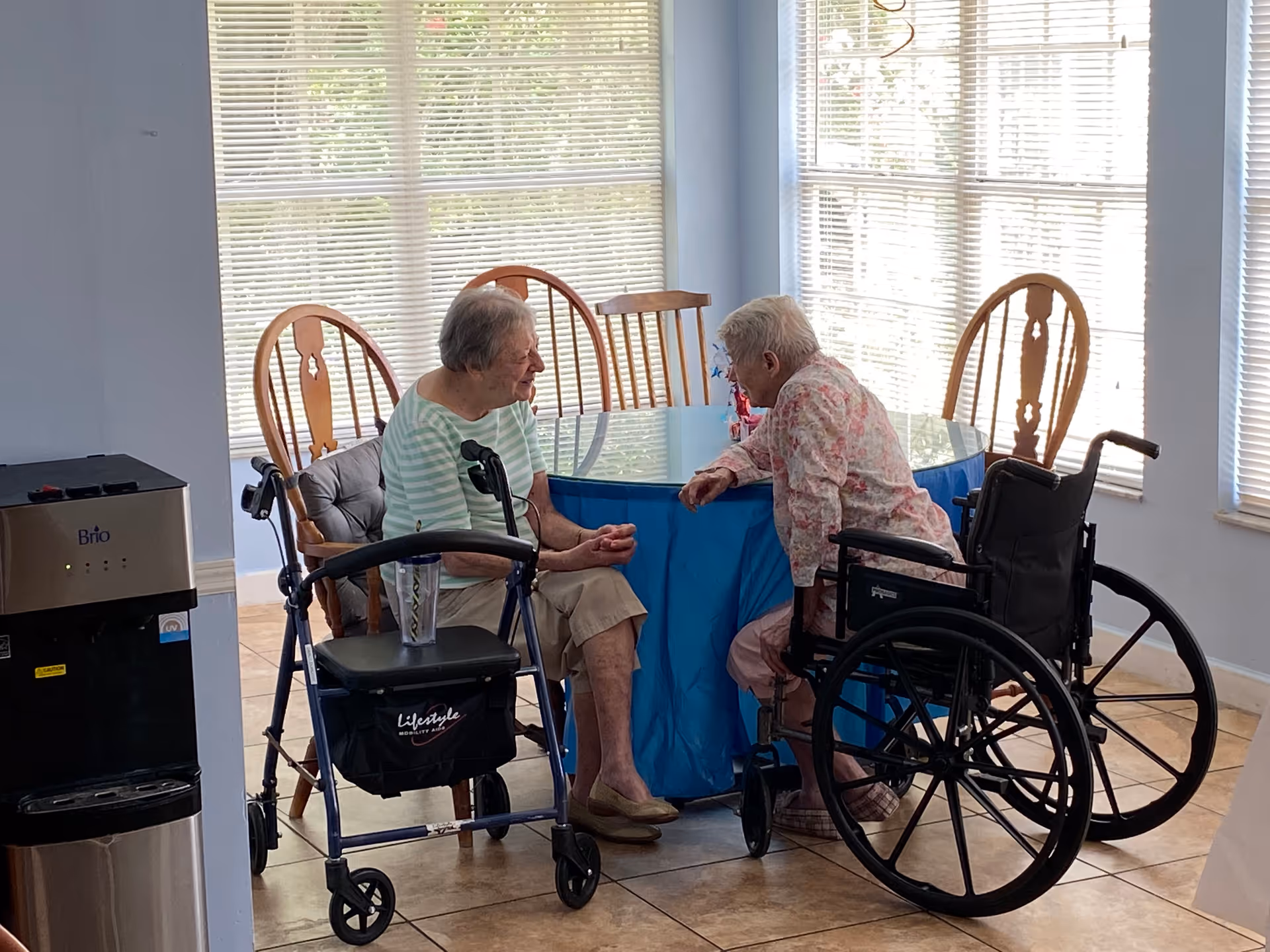 Two elderly women sitting and talking at a round table covered with a blue tablecloth in a bright room with large windows. One woman is seated in a wheelchair and the other on a chair with a walker nearby. There is a water dispenser to the left.