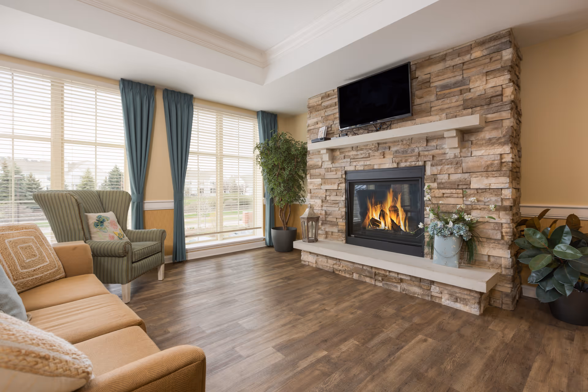 Bright living room featuring a stone fireplace with a mounted TV, sofas and armchairs, large windows with teal curtains, and wood flooring.