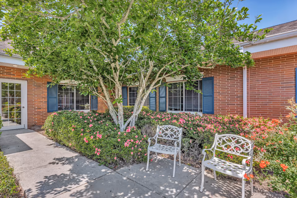 Outdoor patio area with two white metal benches, surrounded by green bushes with pink flowers and a tree. The background shows a brick building with blue window shutters and a glass door.