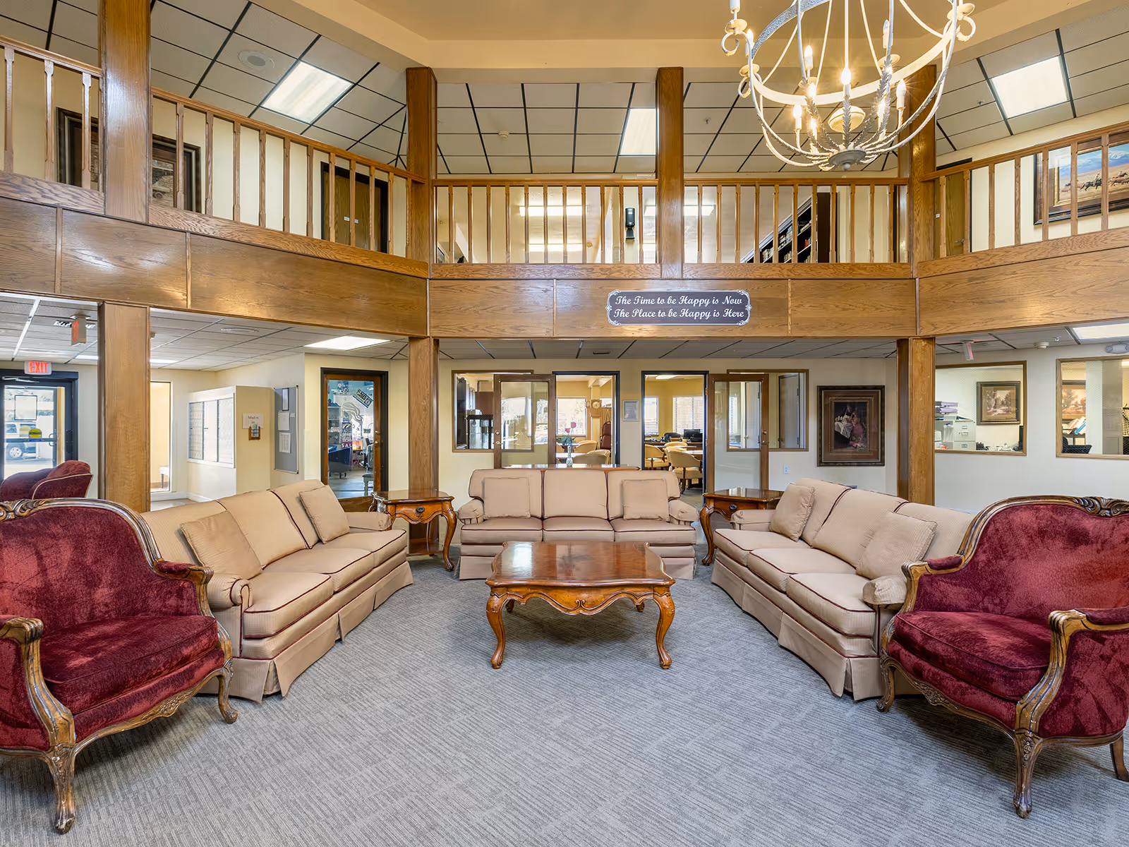 Spacious senior living lobby with beige sofas, red upholstered armchairs, a wooden coffee table, balcony railing and chandelier.