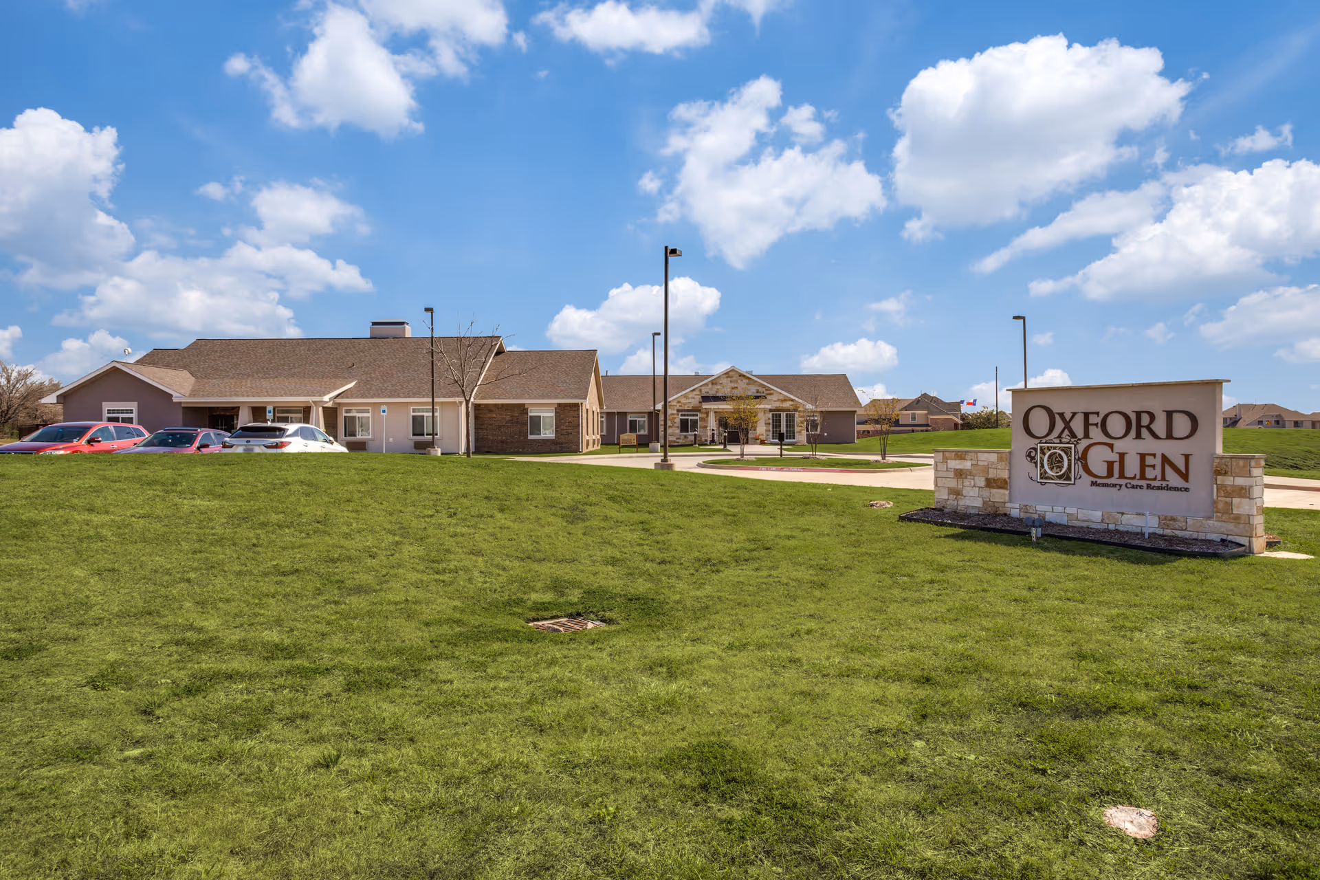 Exterior view of Oxford Glen Memory Care Residence showing a single-story building with a parking area and a large green lawn under a partly cloudy blue sky. A stone sign with the facility name is visible on the right side of the image.