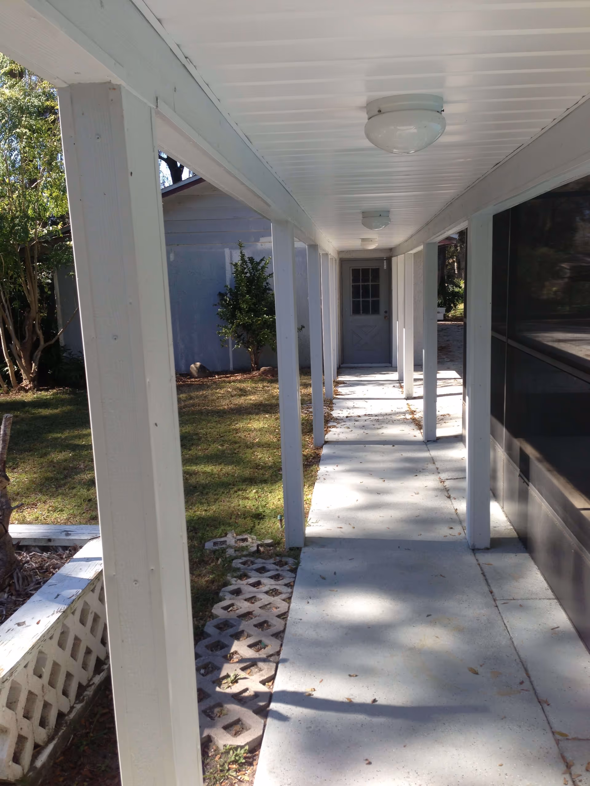 Covered outdoor walkway with white pillars and ceiling lights leading to a closed door at the end. On the left side, there is grass and some trees, and on the right side, there is a dark windowed wall.