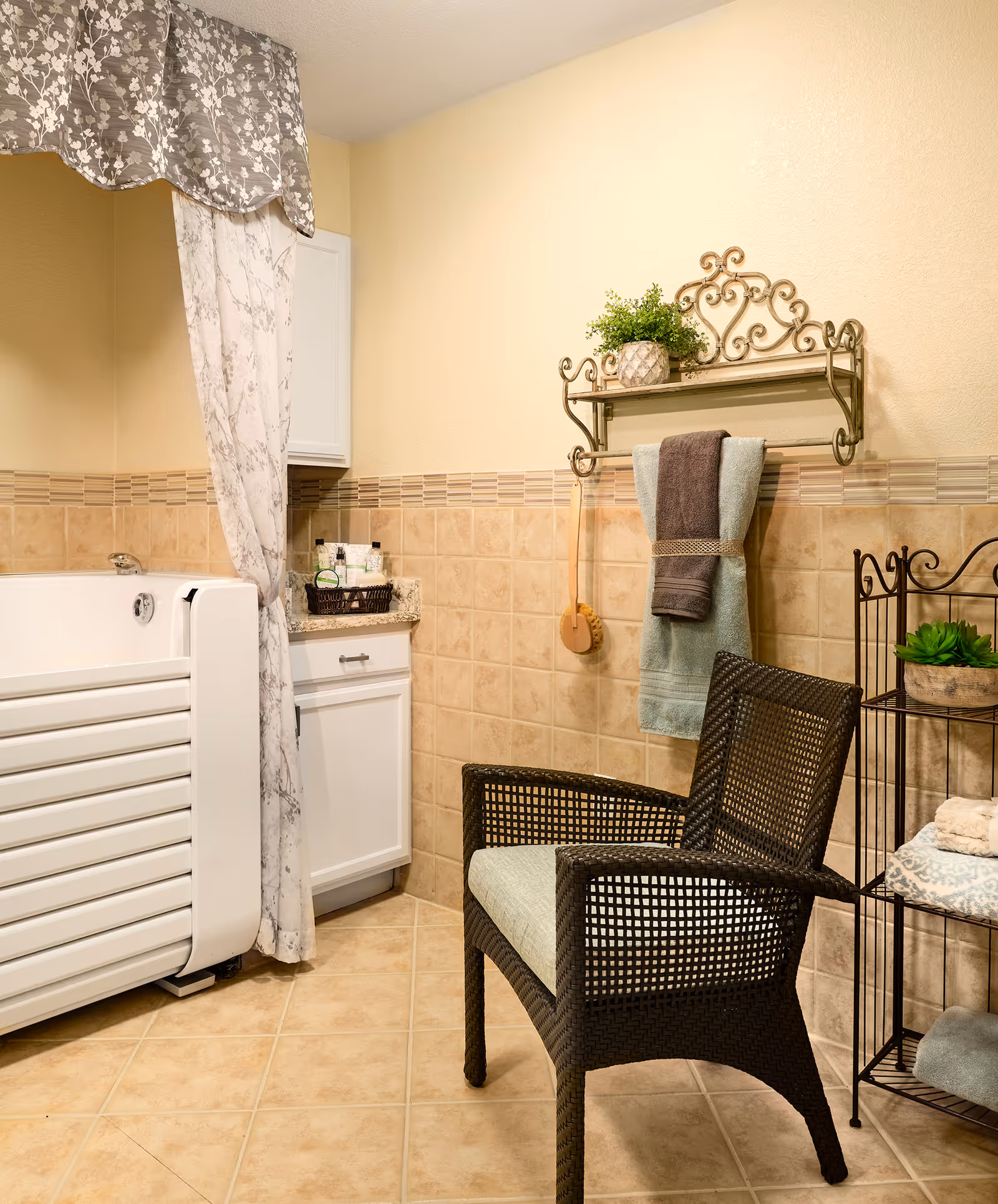 A bathroom with beige tiled walls and floor, featuring a white walk-in bathtub with a floral curtain. There is a white cabinet with toiletries on the countertop, a black wicker chair with a light cushion, a decorative metal wall shelf holding a potted plant and hanging towels, and a black metal shelving unit with folded towels and a small plant.