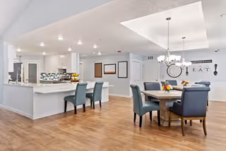 A spacious, well-lit dining area with a wooden floor, featuring a rectangular dining table surrounded by six upholstered chairs in blue and gray tones. To the left is a kitchen counter with two blue chairs, a sink, and a backsplash with a mosaic tile design. The walls are light-colored with decorative elements including a large clock and the word 'EAT' along with utensil wall art. Ceiling lights and a chandelier illuminate the room.