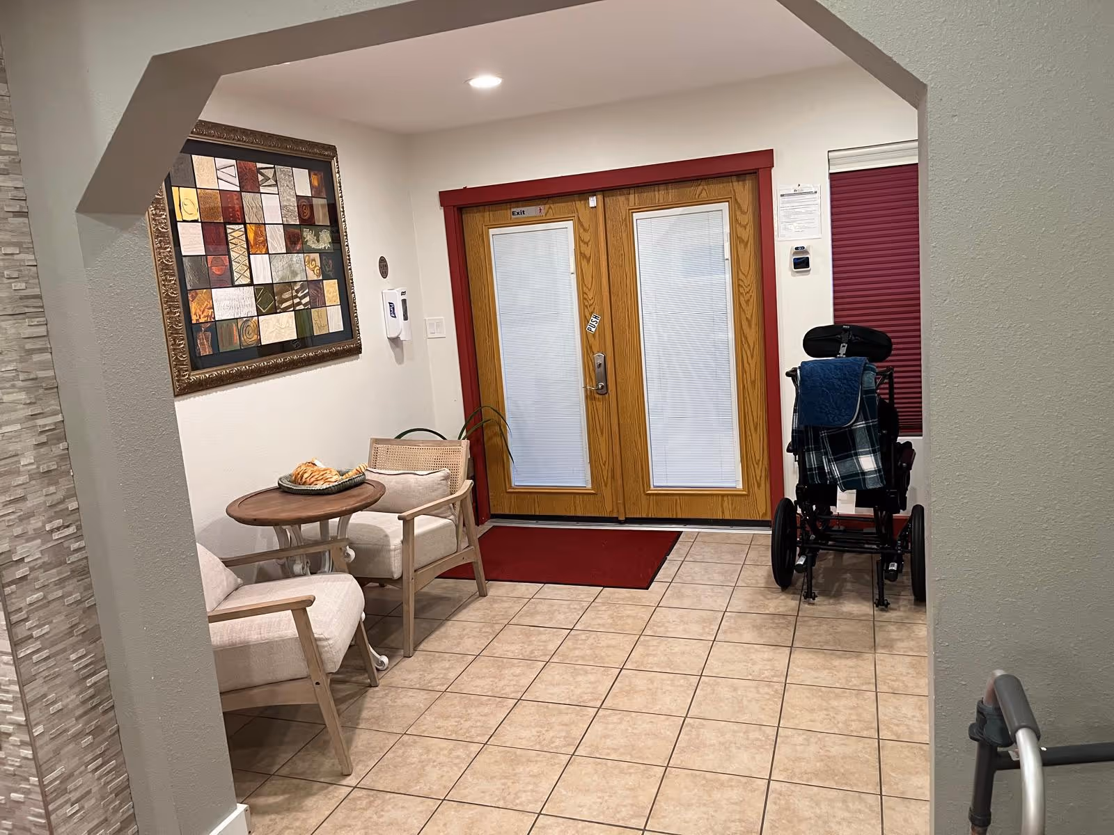 Interior view of a seating area near double wooden doors with glass panels covered by blinds. The area has tiled flooring, two cushioned chairs around a small round wooden table with a decorative item on it, a framed abstract artwork on the wall, a red floor mat in front of the doors, and a wheelchair with a plaid blanket draped over it. The walls are painted white with a stone accent on one side.