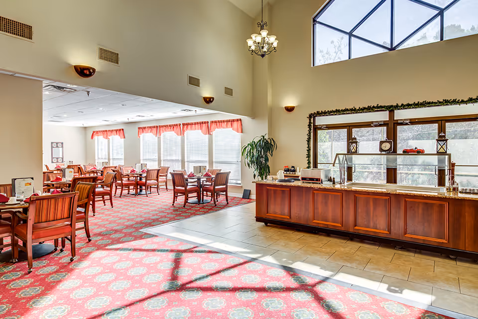 Bright dining room area with multiple wooden tables and chairs arranged on a red patterned carpet. Large windows with red valances allow natural light to fill the space. A wooden buffet counter with a glass sneeze guard is positioned near the entrance, decorated with small lanterns and a clock. High ceilings with a chandelier and wall sconces add to the spacious feel.