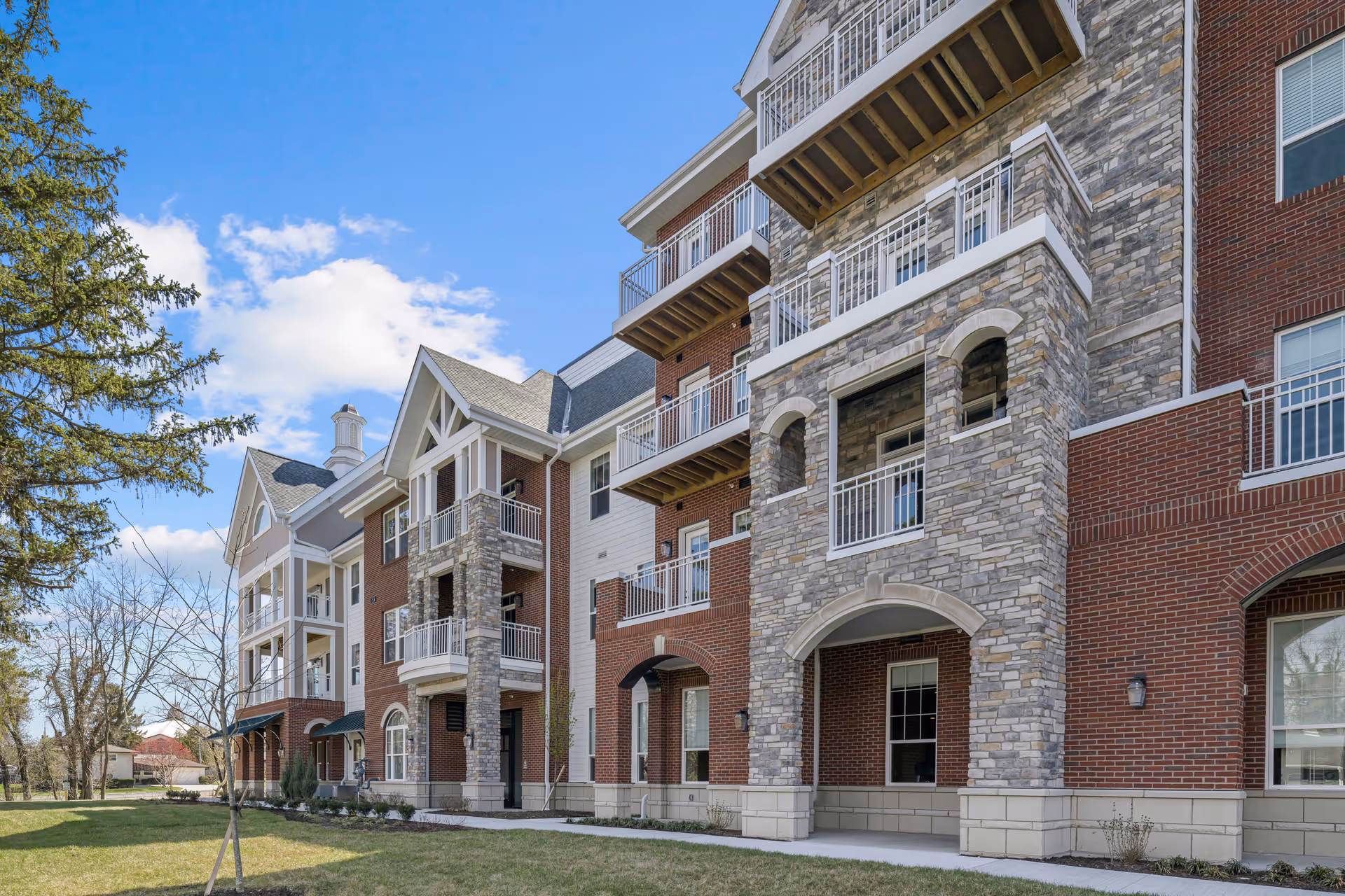 Front exterior of a multi-story brick and stone residential building with balconies, lawn, and trees under a blue sky.