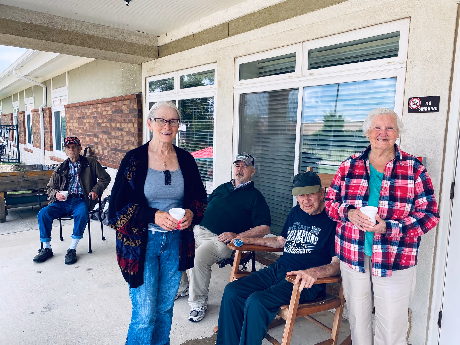 Five elderly individuals sitting and standing on a covered outdoor patio area of a building. Two women are standing in the foreground holding white cups, while three men are seated behind them. The building has large windows with blinds and a 'No Smoking' sign on the wall.