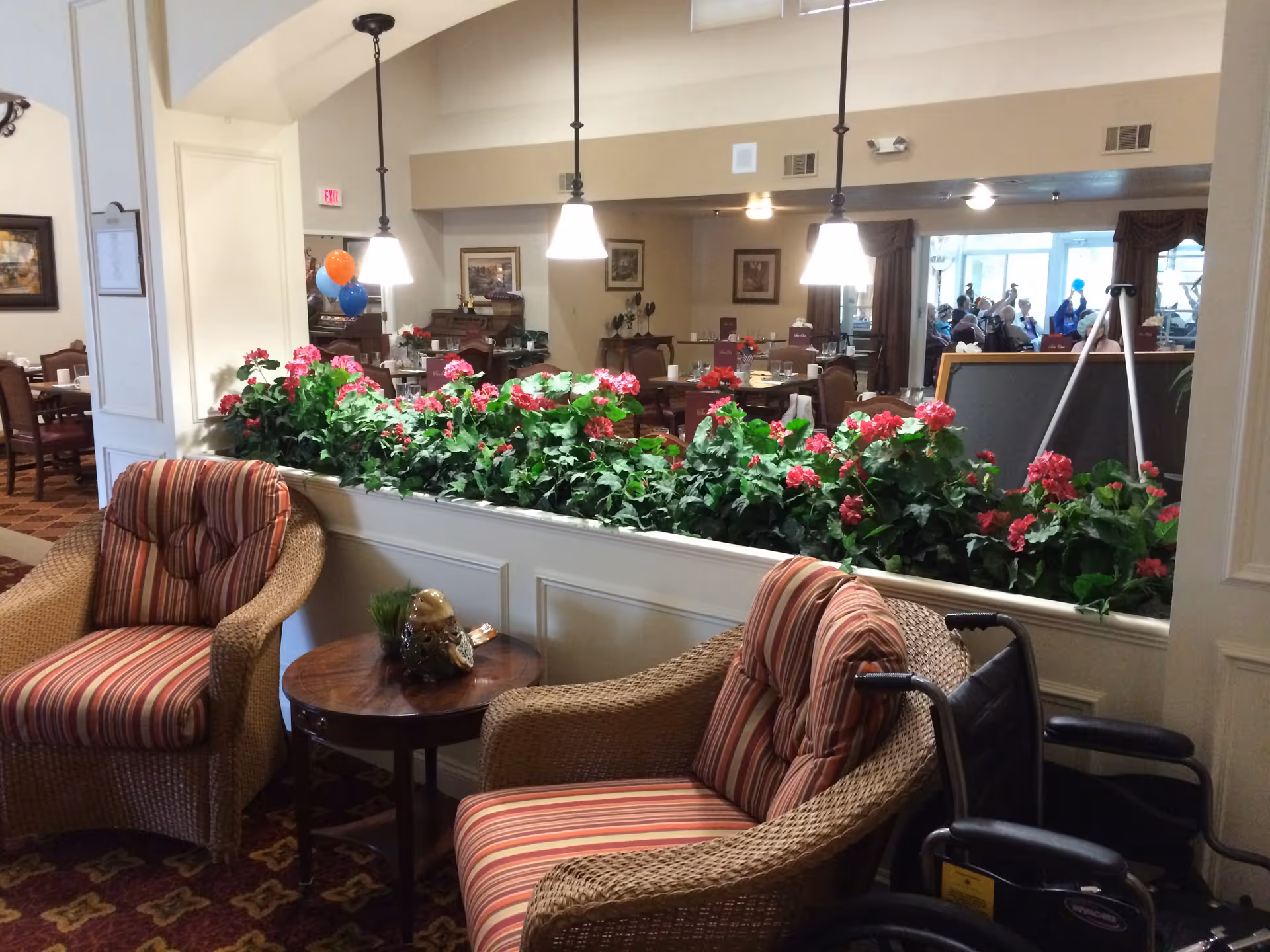 Cozy assisted-living dining/lounge area with wicker armchairs, striped cushions, a long planter of pink flowers, and dining tables with pendant lights in the background.
