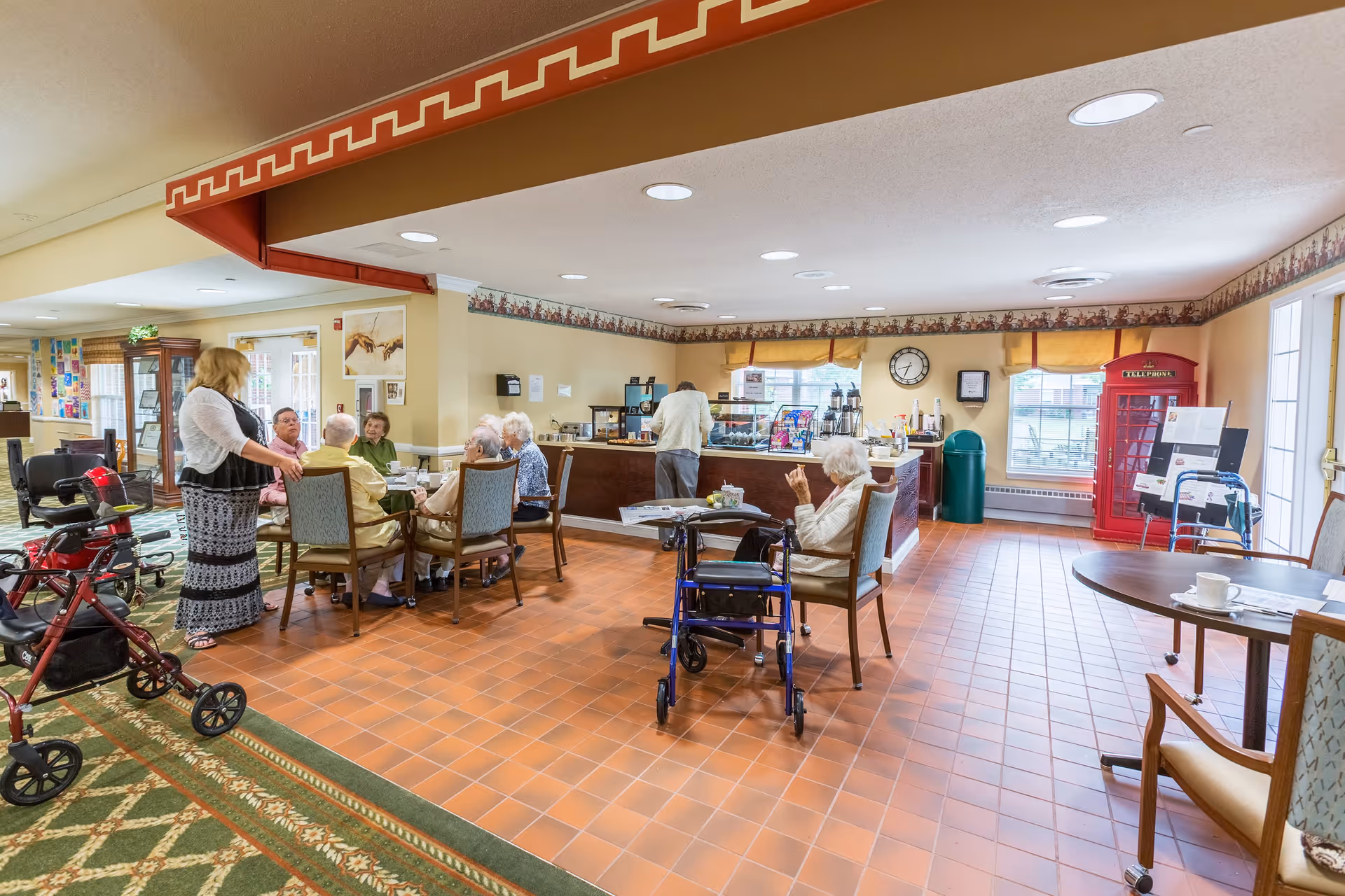 A common area in a senior living facility with elderly residents sitting around tables and engaging in conversation. One person is standing and talking to the group. The room has tiled flooring, a carpeted section, and a counter with coffee and snacks. There is a red British-style telephone booth in the corner near windows, and mobility aids like walkers are visible.