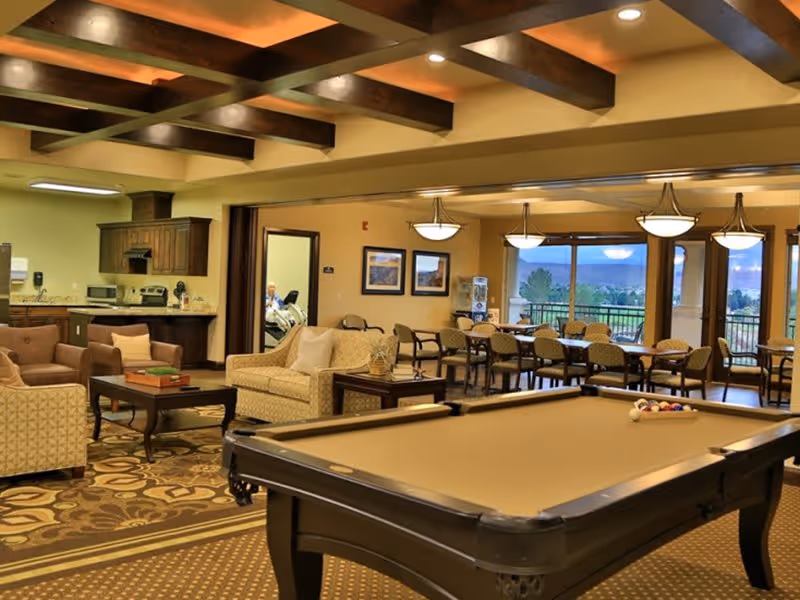 A spacious common area in a senior living facility featuring a pool table in the foreground, comfortable seating with sofas and armchairs, a small kitchen area with appliances, and a dining area with tables and chairs near large windows offering a scenic view outside.