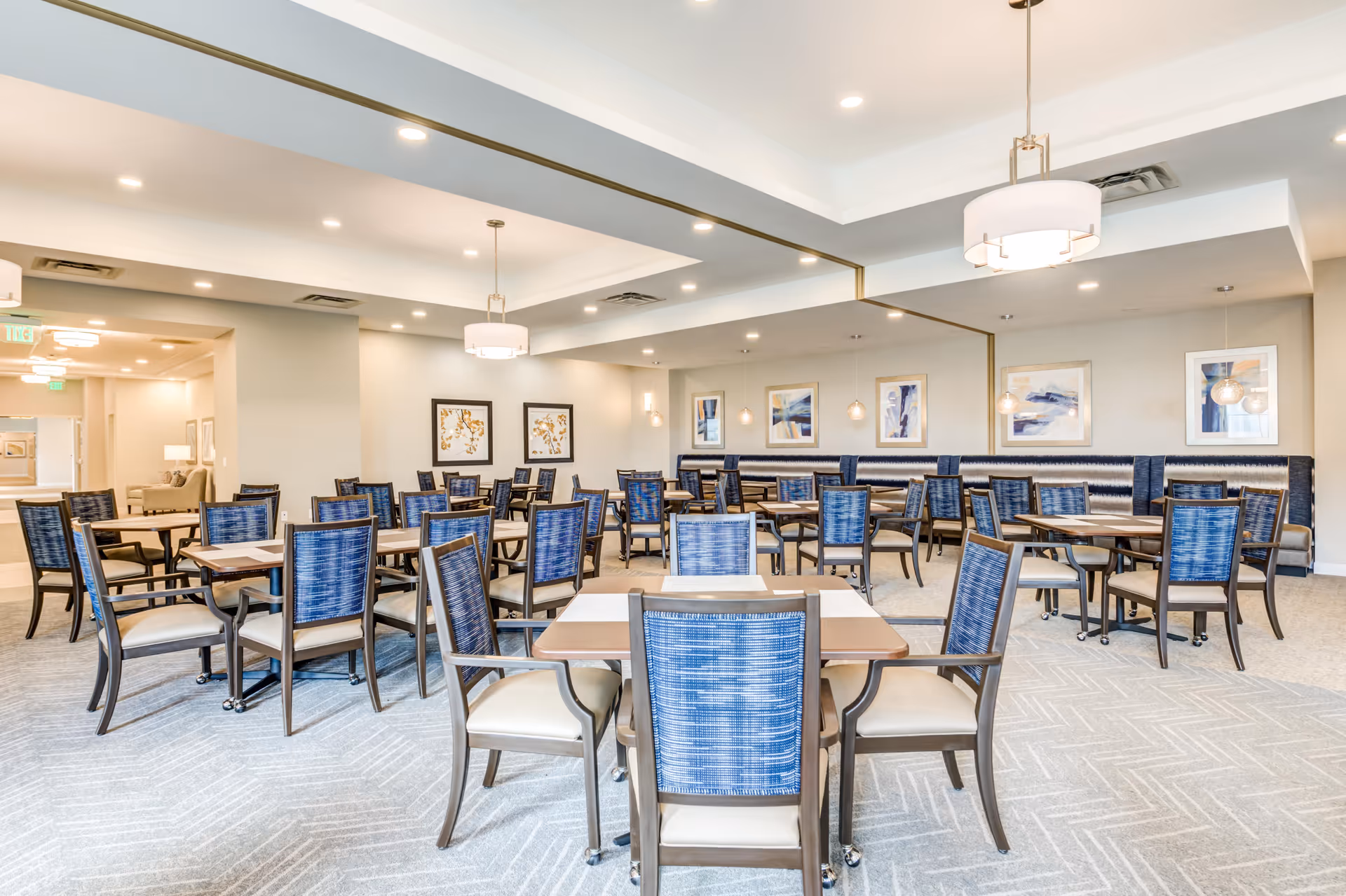 A spacious and well-lit dining room with multiple tables and chairs arranged neatly. The chairs have blue patterned backs and beige seats. The room features a large mirror on one wall, modern light fixtures hanging from the ceiling, and framed artwork on the walls. The carpet is light-colored with a subtle pattern.
