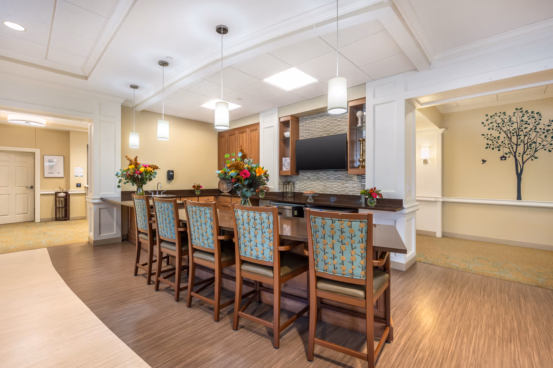 Interior view of a senior living facility's common area featuring a long counter with six wooden chairs upholstered in blue patterned fabric. The counter has flower arrangements and pendant lights hanging above. In the background, there is a wall-mounted TV and cabinetry with a tiled backsplash. The room has light-colored walls and flooring, with a decorative tree wall decal visible in an adjacent hallway.