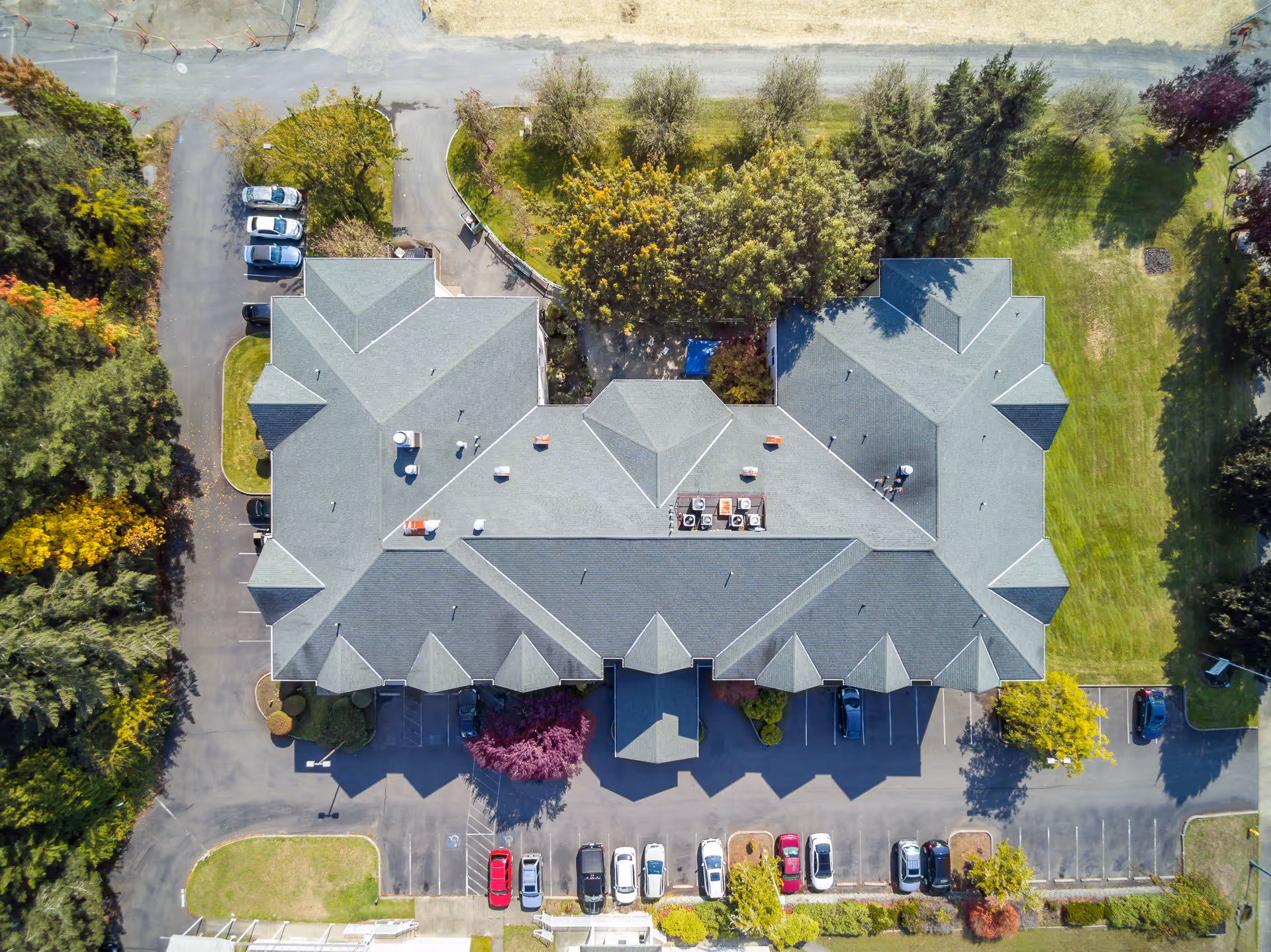 Aerial view of a large building with a gray roof surrounded by parking lots and greenery. Several cars are parked in the parking areas around the building, and trees and grass are visible on the property.
